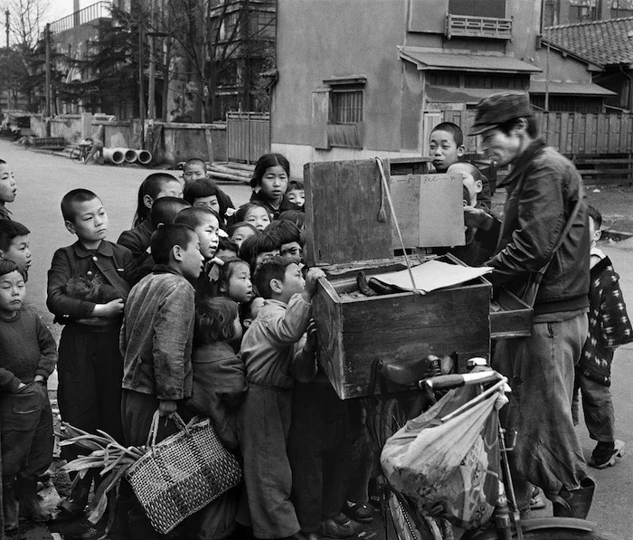 Historische foto: een kamishibai-verteller met zijn fiets en houten theatertje, omringd door kinderen in Japan